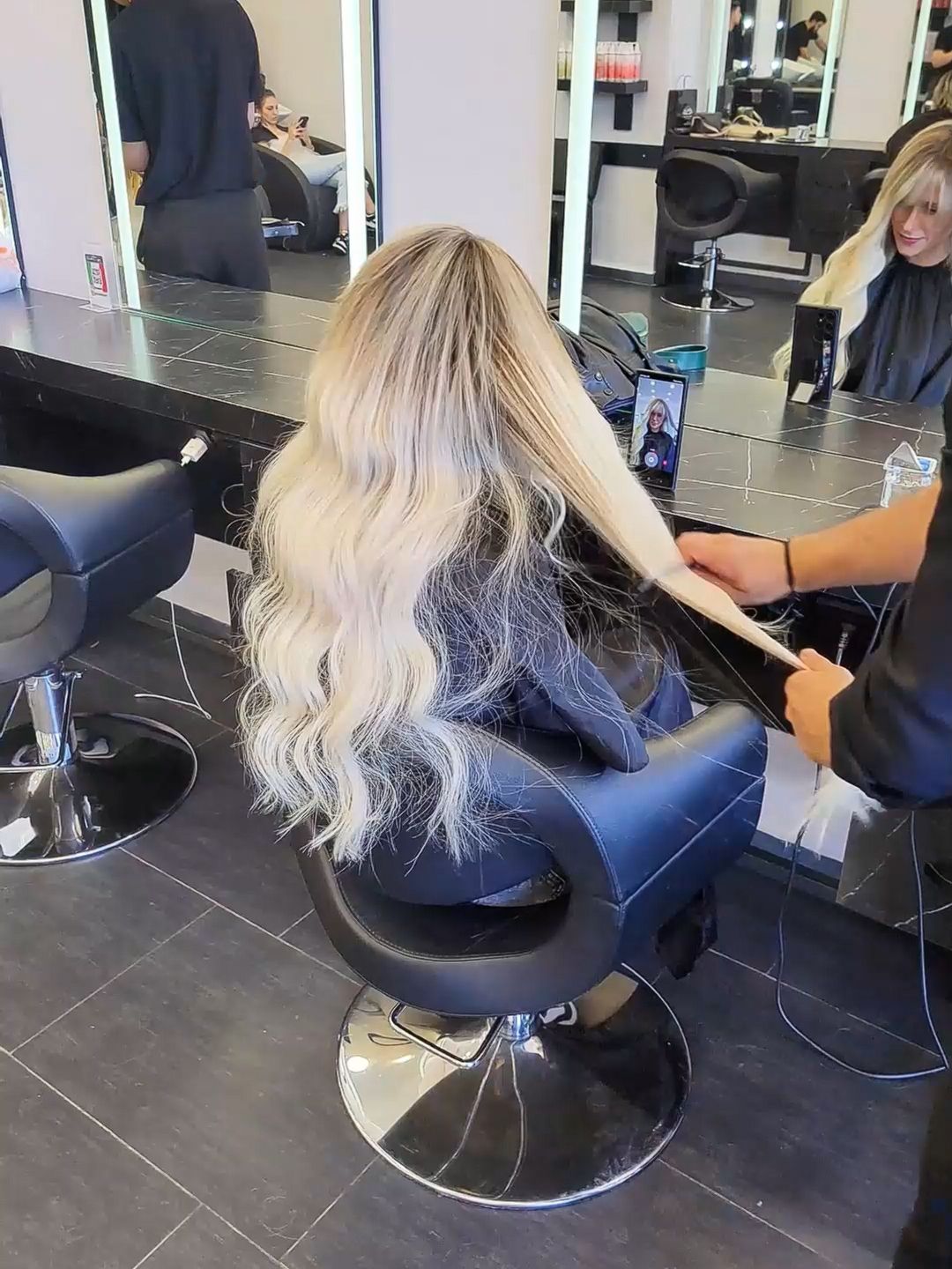 Blonde woman sitting in a salon chair with long wavy hair as stylist prepares her hair during beauty appointment.