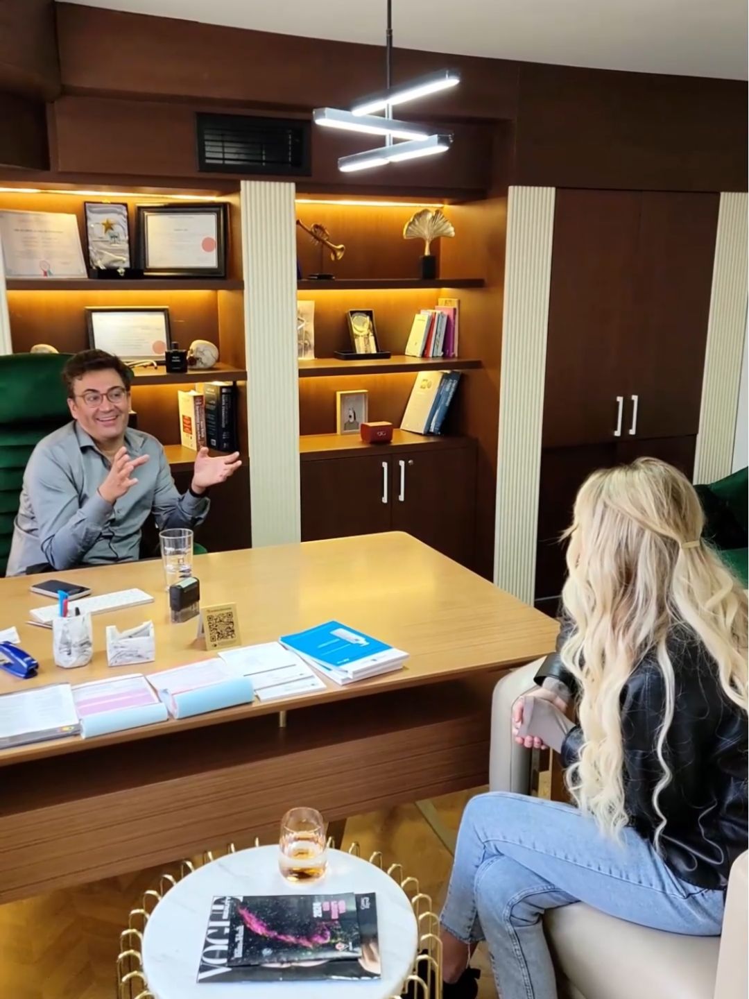 Blonde woman sitting across a desk consulting with a cosmetic doctor in a well-decorated office with certificates.