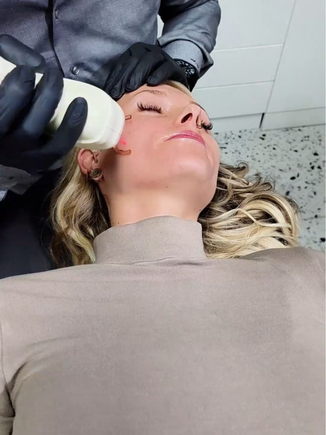 Blonde woman receiving a laser facial treatment from a professional wearing gloves, focusing on cheek area in clinic.
