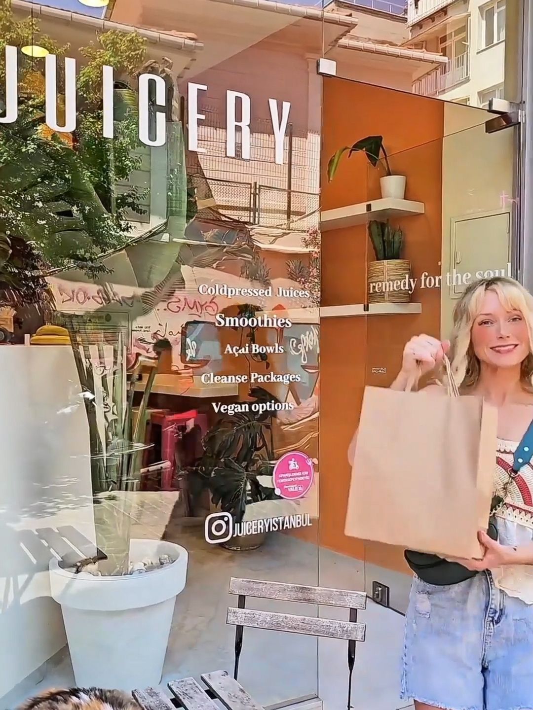 A woman smiling with a shopping bag in front of Juicery Istanbul, with the shop’s glass window and cat on a wooden bench.