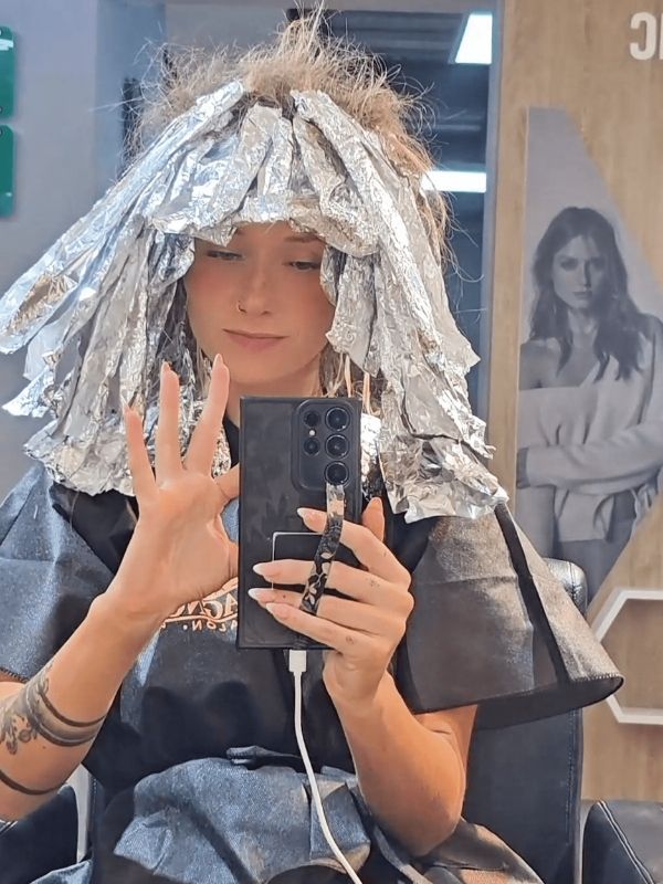 A woman with foil in her hair takes a mirror selfie during a blonding and color correction process at a salon.