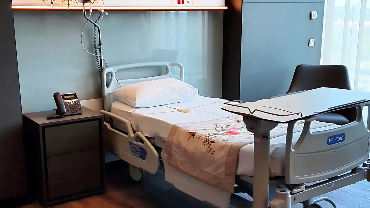 A modern hospital patient room at Memorial Hospital in Istanbul, featuring a single bed, bedside table with phone, and large window with natural light.