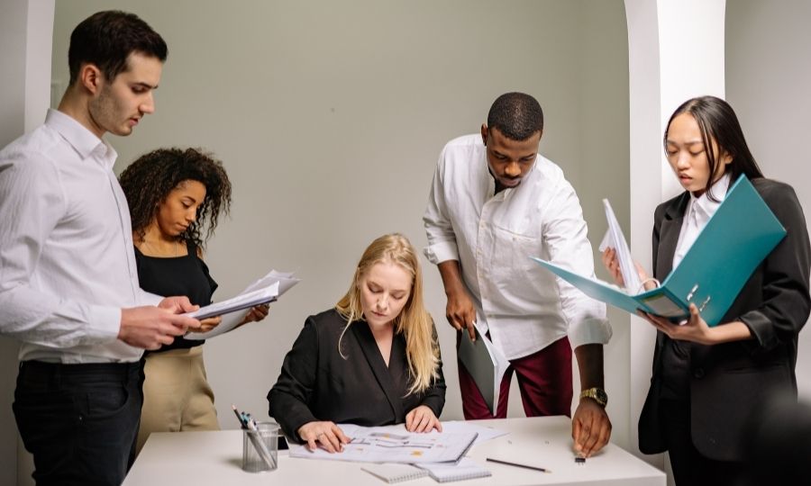  Five professionals collaborating in a modern office, reviewing documents and a teal folder.