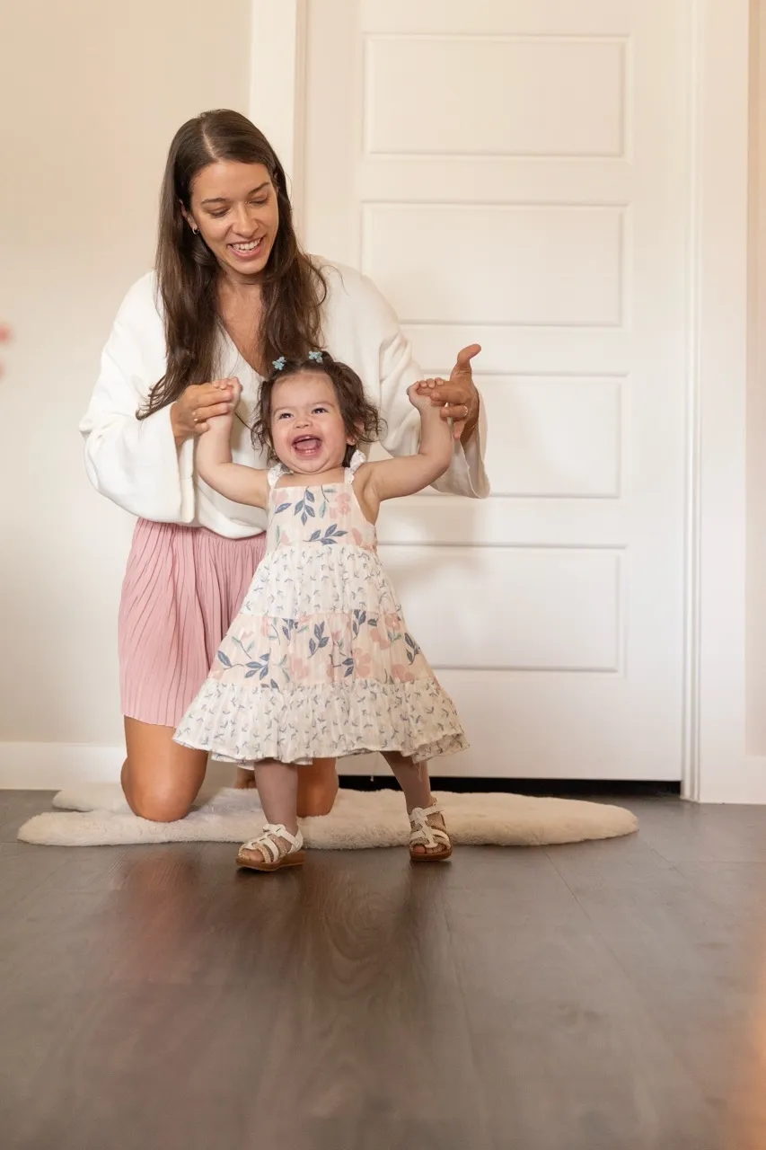 Smiling toddler in a floral dress taking steps while holding hands of a kneeling woman.