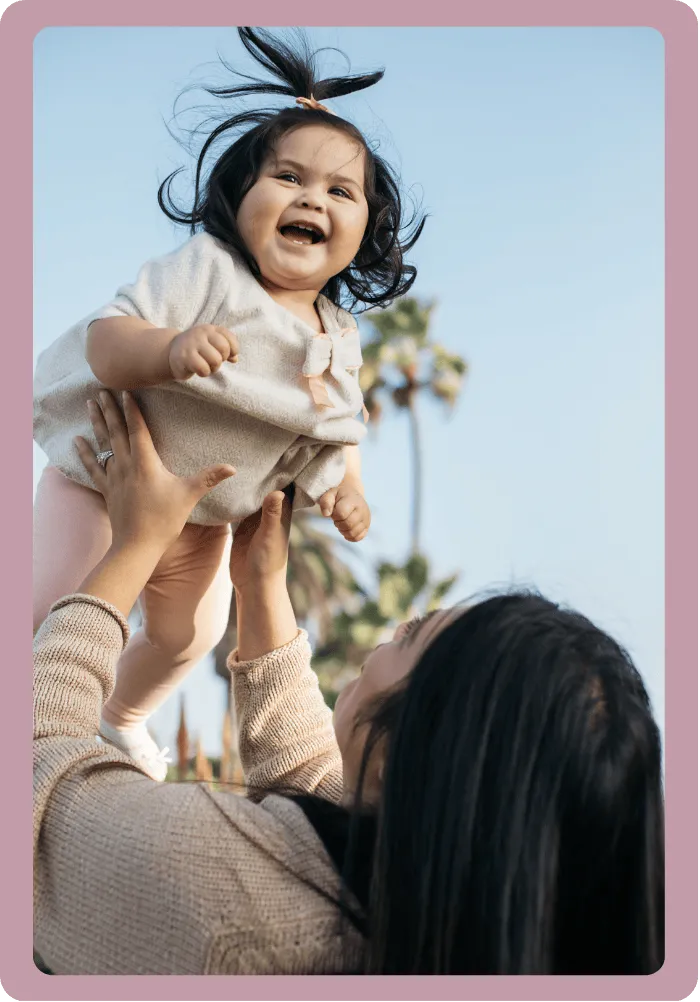 Smiling baby lifted in the air by a woman with long dark hair outdoors, with palm trees and blue sky in the background.