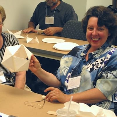 Two middle aged adults smiling, holding up their paper folded sculptures during a wholemovement workshop.