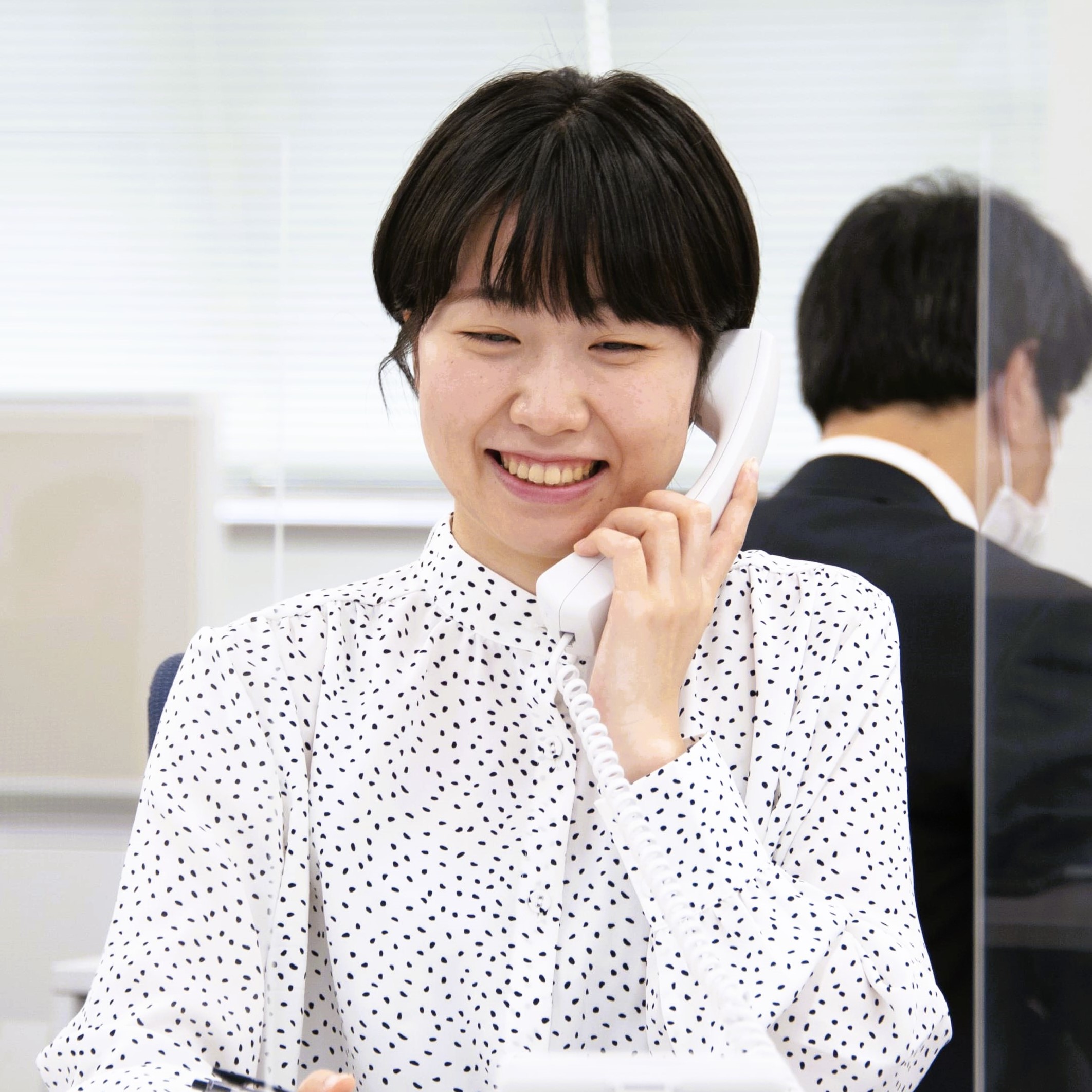 Smiling woman in a white shirt with black dots talking on a white office telephone.