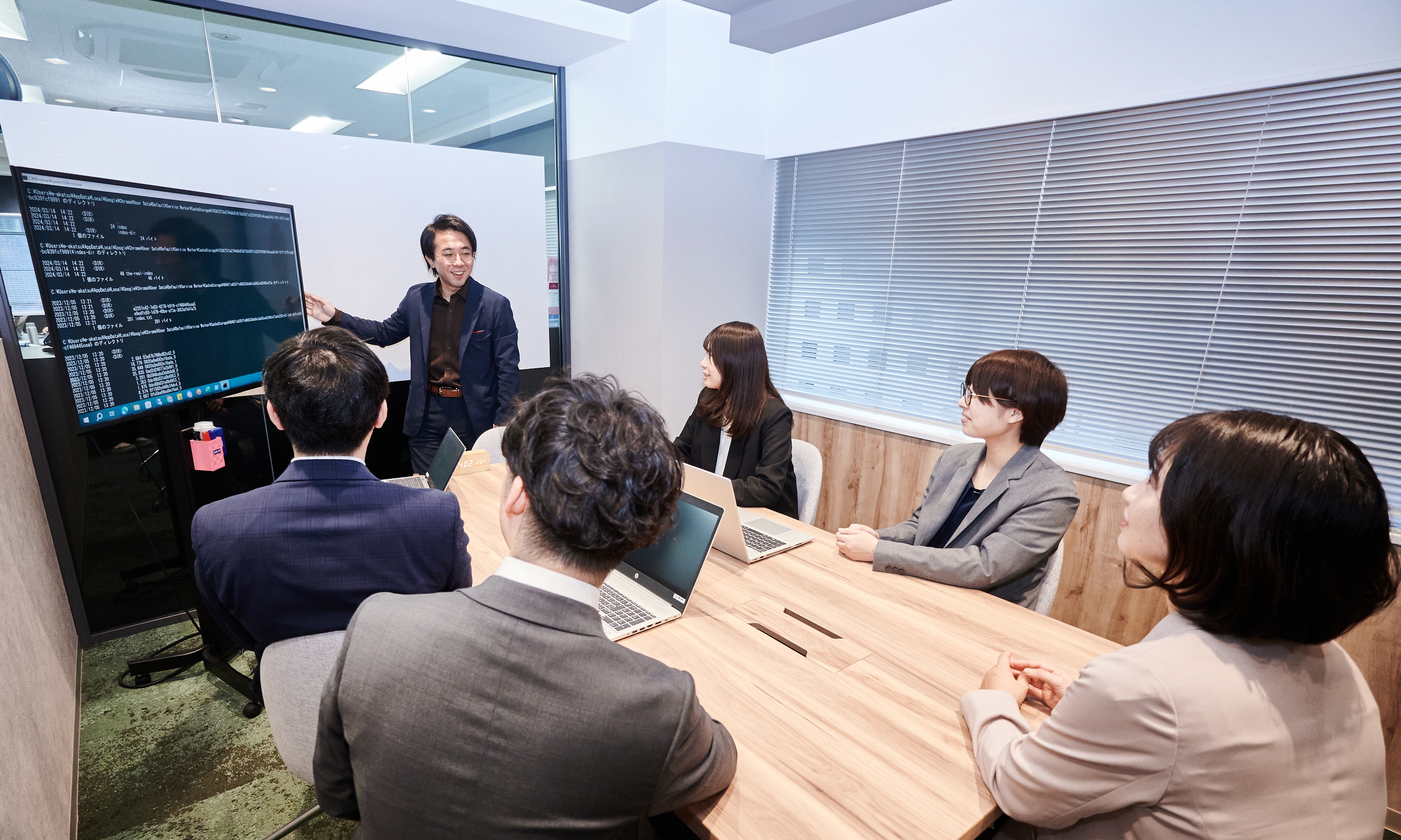 Man in business attire presenting programming code on a large screen to a group of five attentive colleagues seated around a wooden conference table in a modern office.