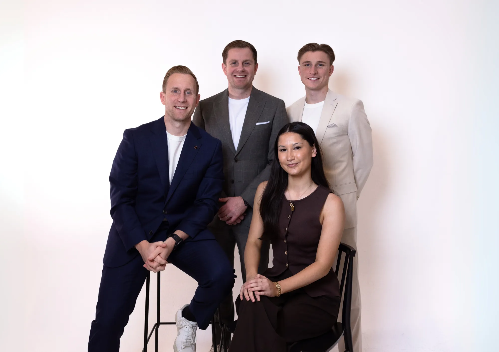 Group photo of three men in suits and a woman in a dark sleeveless dress, all smiling against a white background.