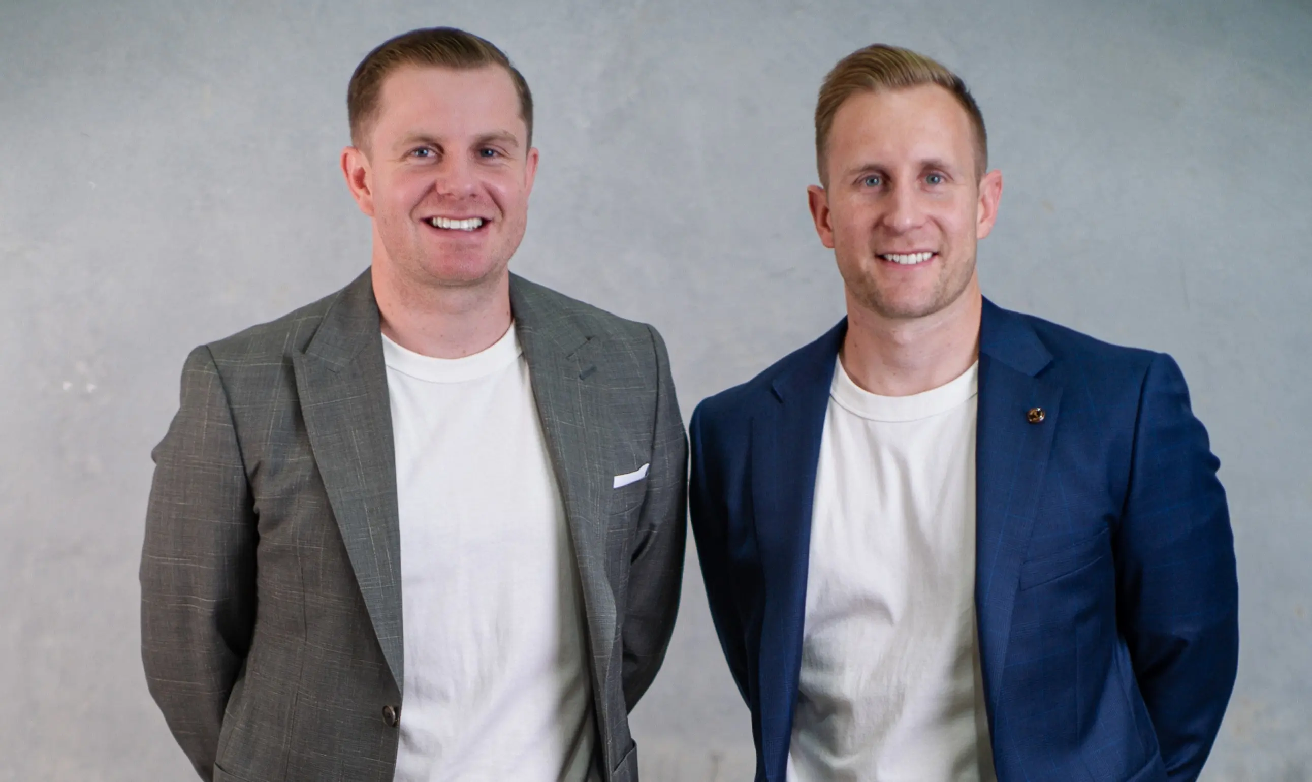 Two men smiling and standing side by side wearing white t-shirts with blazers, one gray and one navy blue, against a gray background.