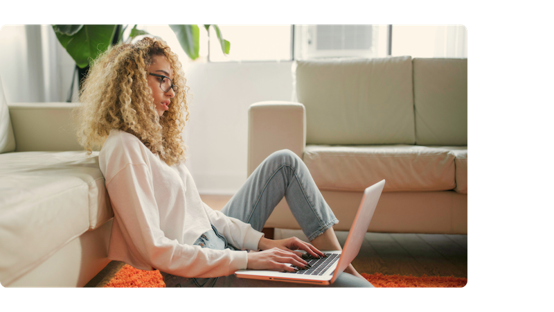 A woman with curly blonde hair and glasses sits on the floor against a sofa, working on a laptop in a bright, relaxed living room setting.