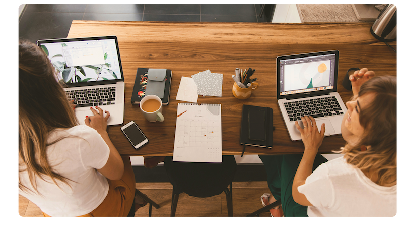 Two women work side by side at a wooden table, each using a laptop, with notebooks, a calendar, and coffee cups spread between them, suggesting a collaborative or remote work setting.