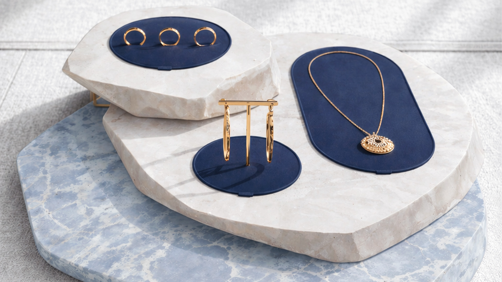 De Beers gold jewelry displayed on blue velvet pads over sculptural marble plinths.