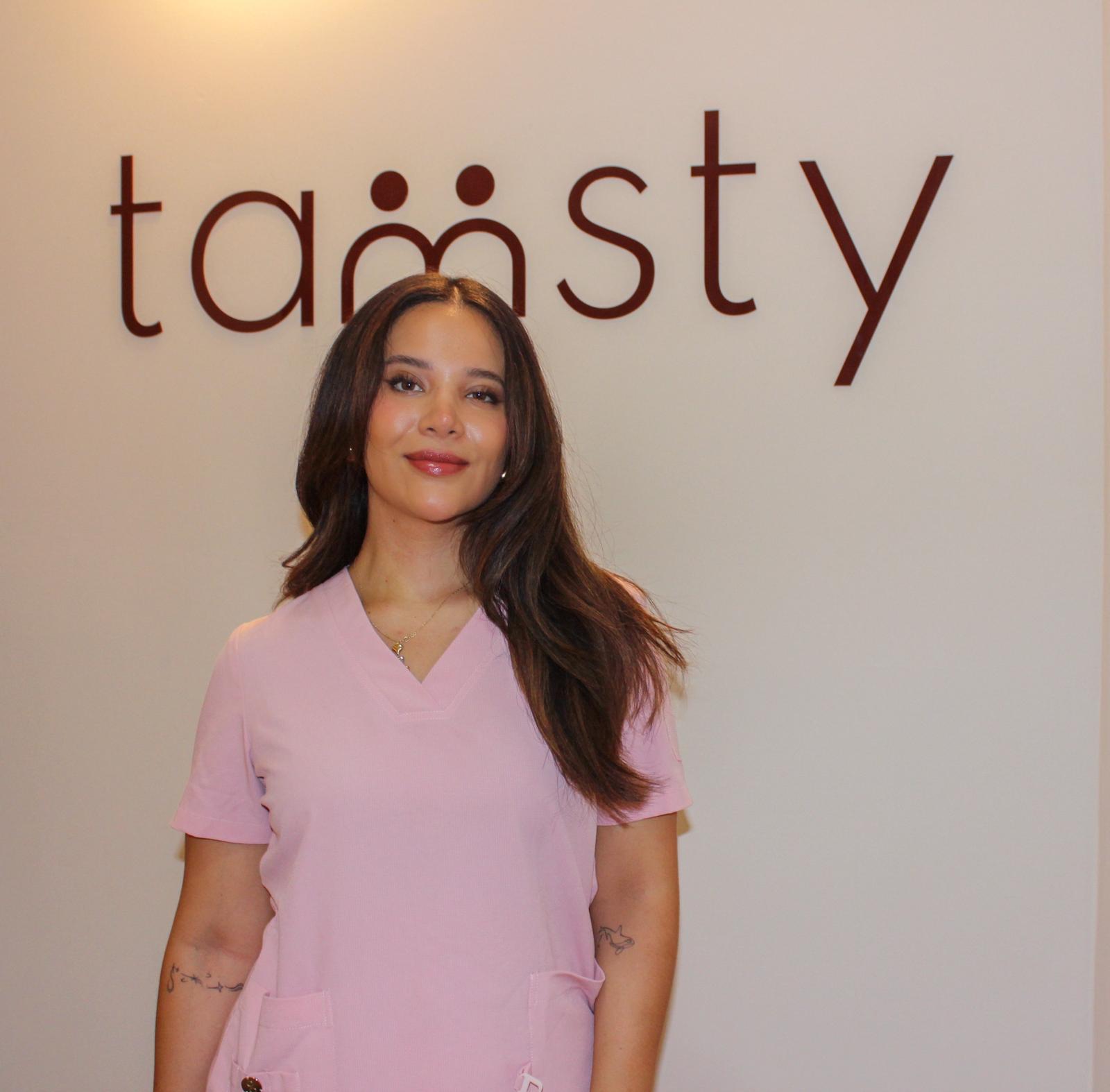 Tamsty Suárez, nutricionista clínica y experta en bienestar digestivo, frente al logo de su marca, con uniforme rosa y expresión serena, representando enfoque integral y cercano en salud.