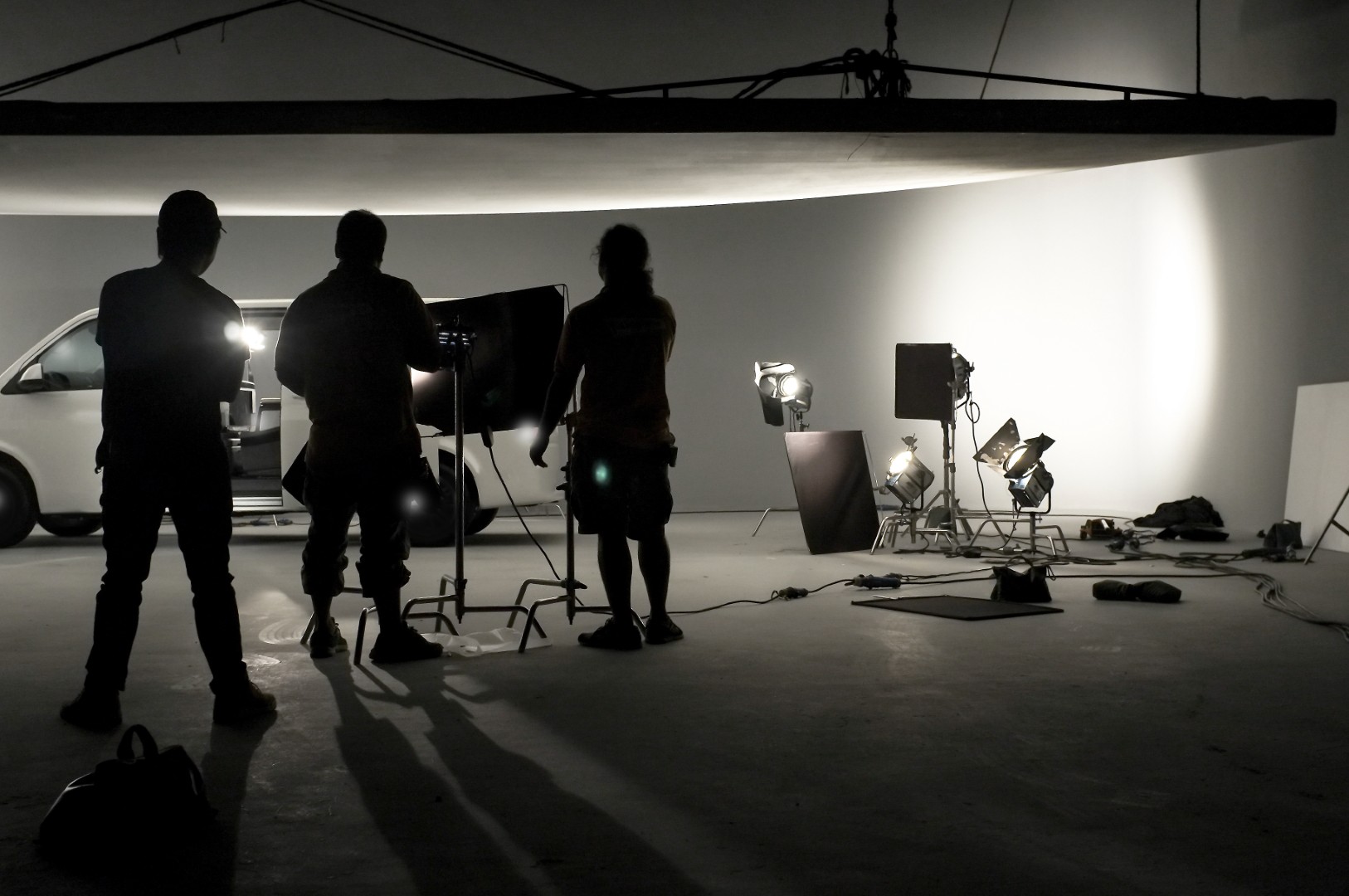 Silhouettes of three people standing in a dimly lit studio with lighting equipment and a white van in the background.