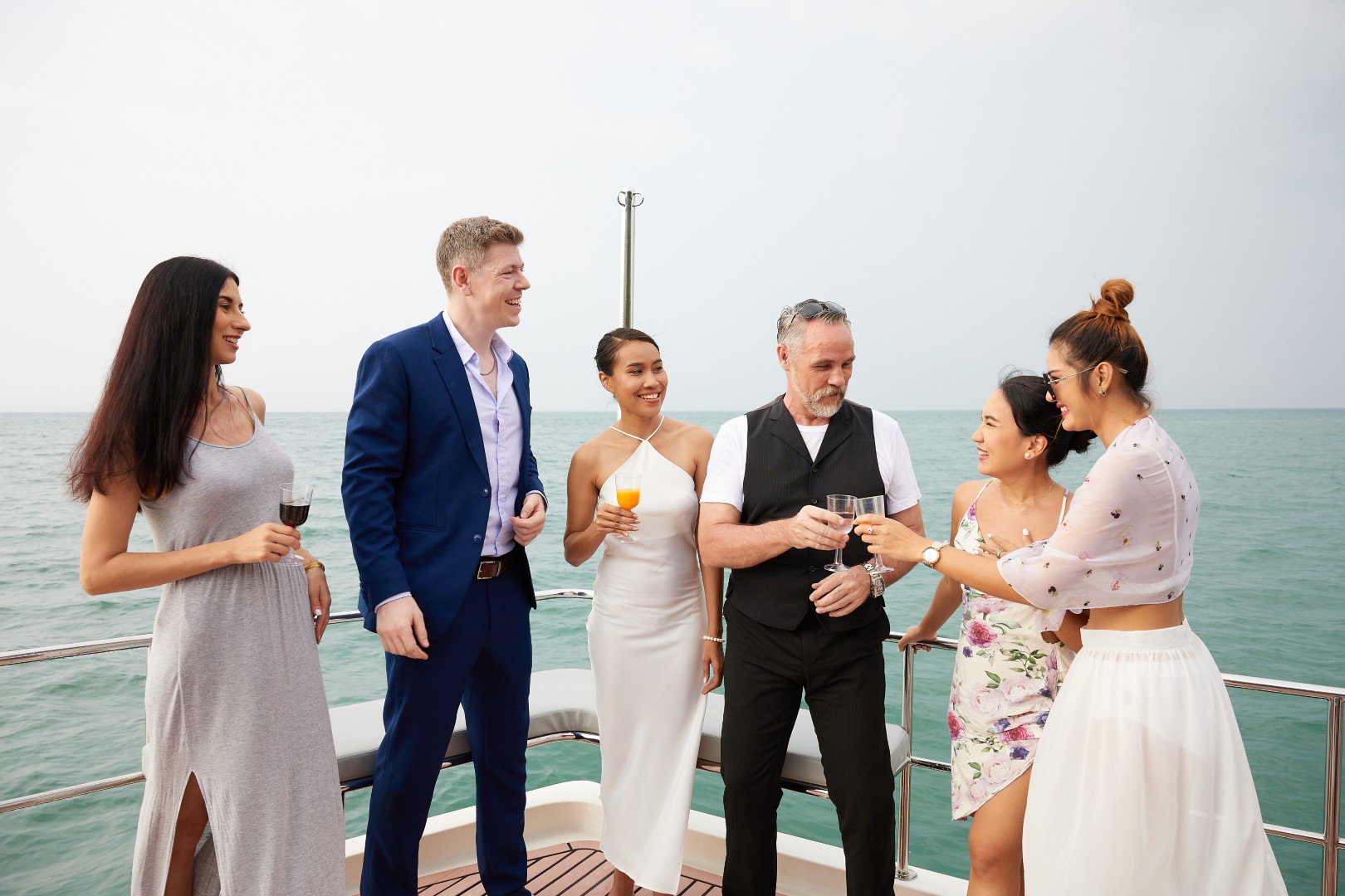 Group of three women and two men dressed elegantly, smiling and holding drinks on a boat with the ocean in the background.