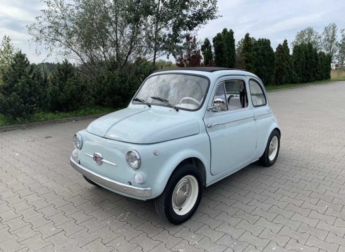 Light blue vintage Fiat 500 parked on a paved area near green trees and bushes.