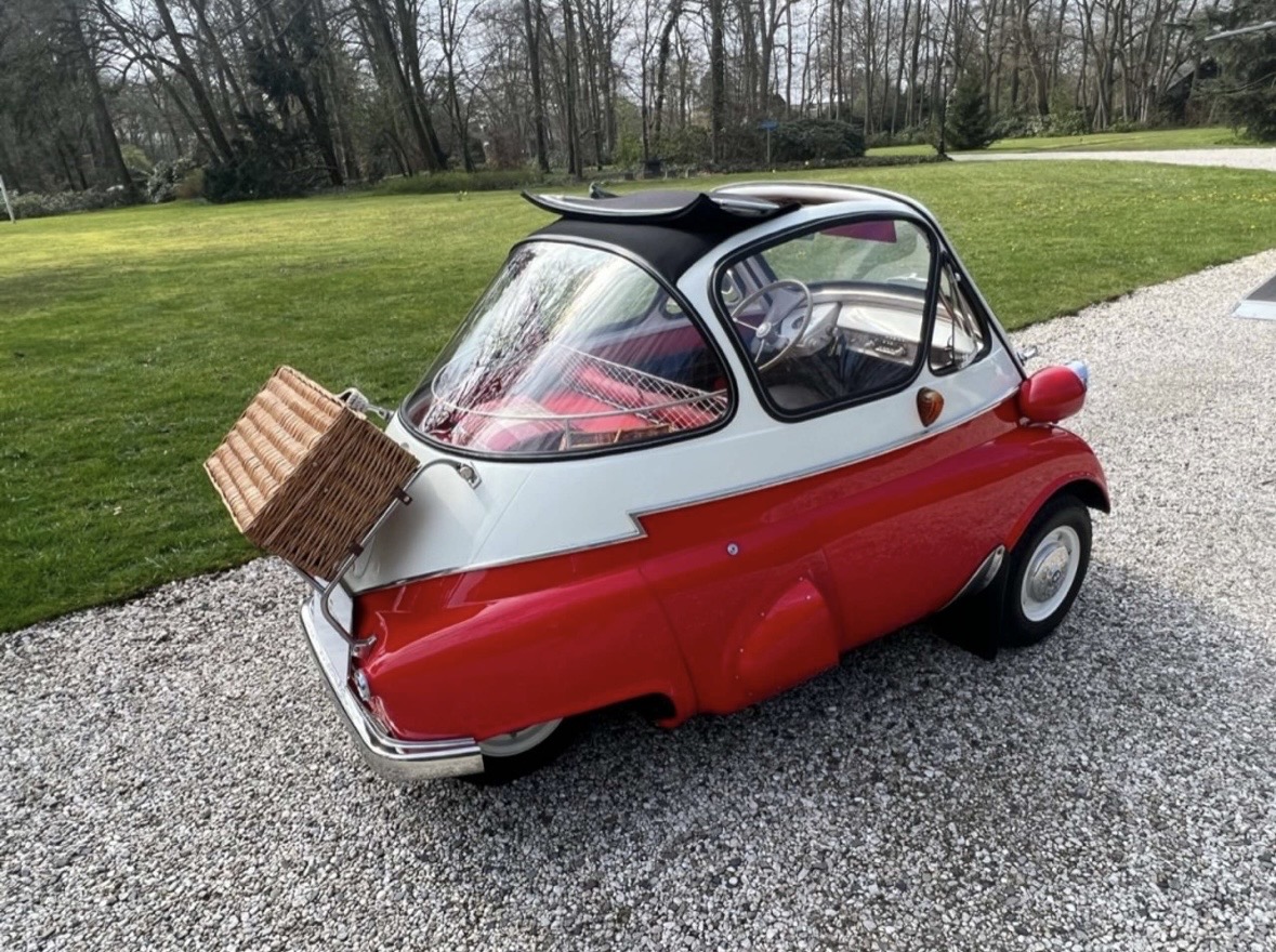 Red and white vintage BMW Isetta microcar with a wicker picnic basket mounted on the rear, parked on a gravel driveway near a grassy area.