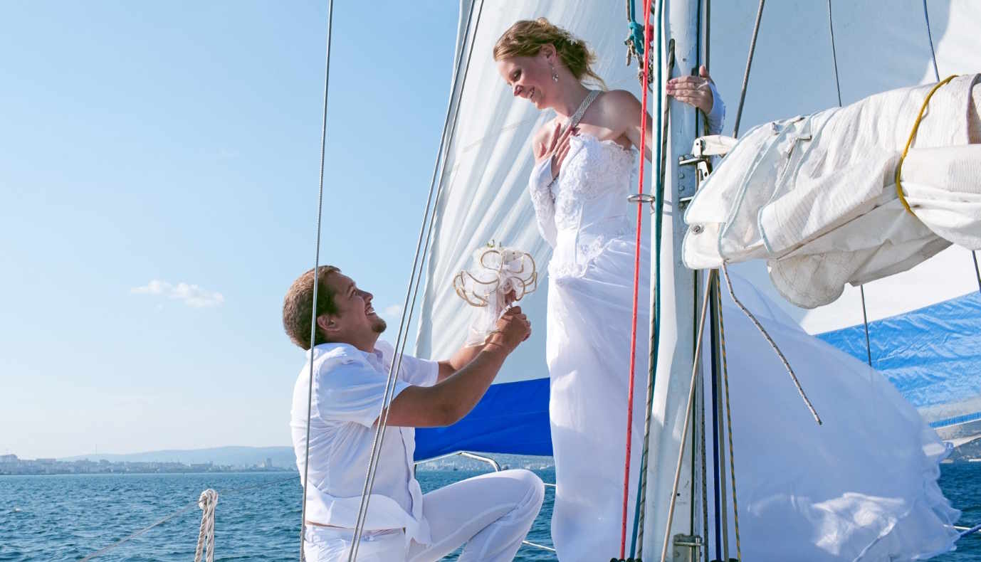 Man in white outfit kneeling and holding a bouquet, proposing to a woman in a white wedding dress on a sailboat.