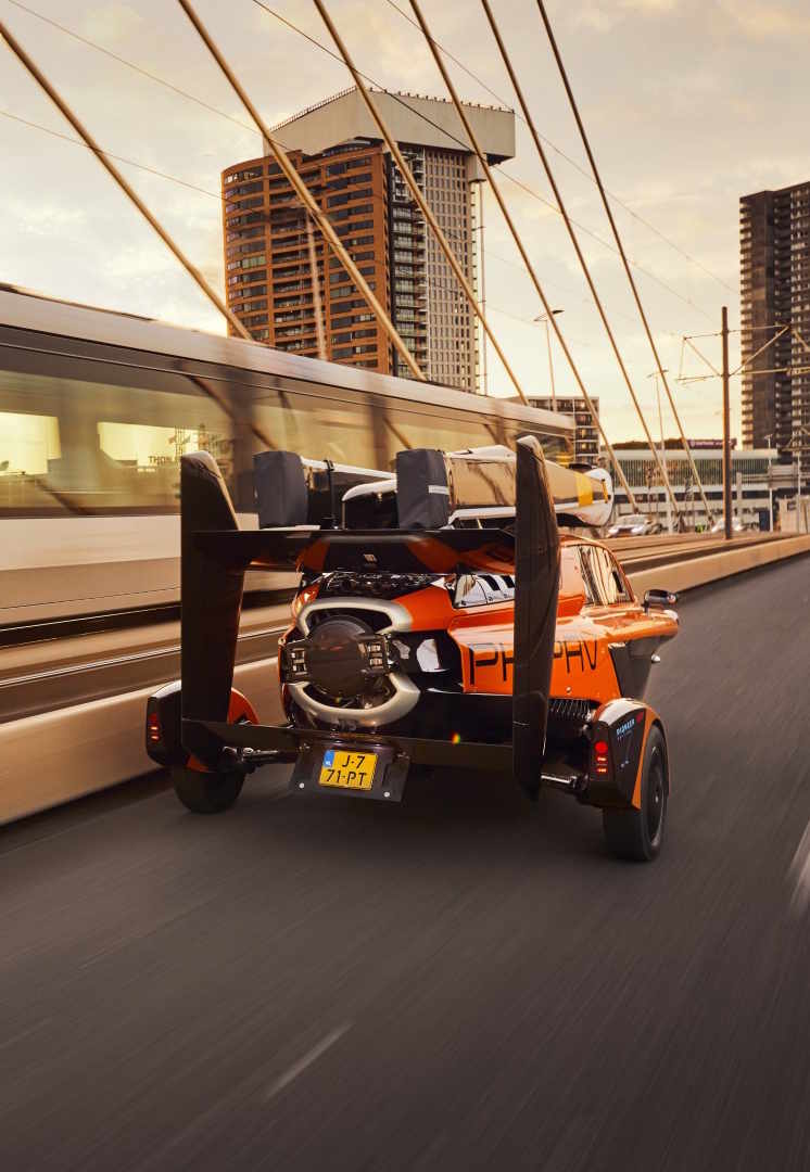 Rear view of an orange and black prototype flying vehicle on a city bridge with a moving train and tall buildings in the background.
