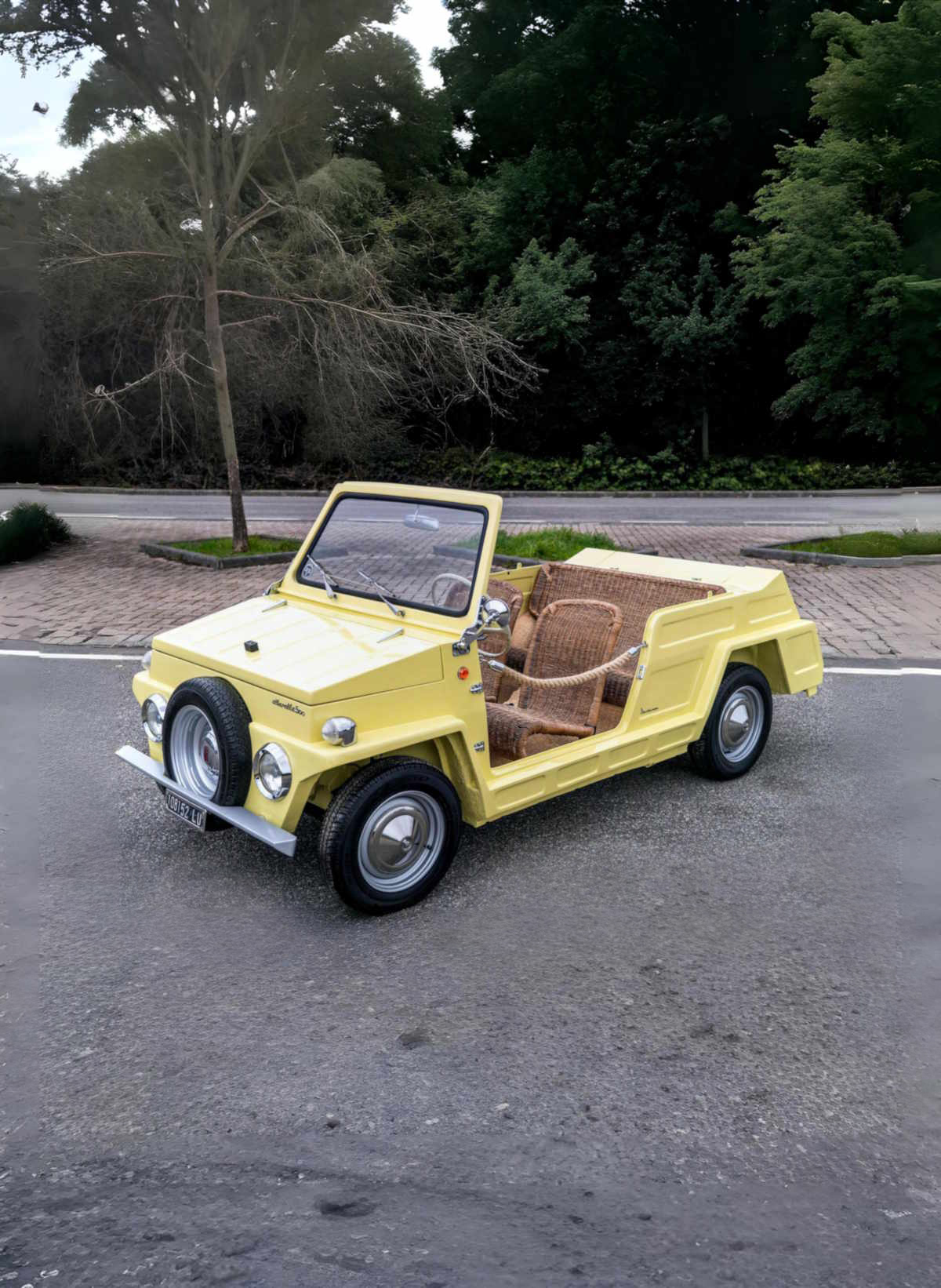 Yellow vintage open-top car with wicker seats parked on a paved road lined with trees and greenery.