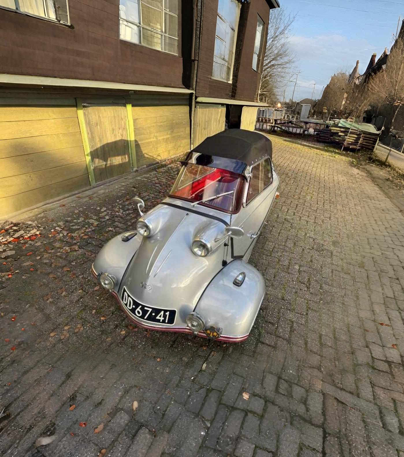 Silver vintage three-wheeled car with a black soft top parked on a cobblestone street beside yellow garage doors.