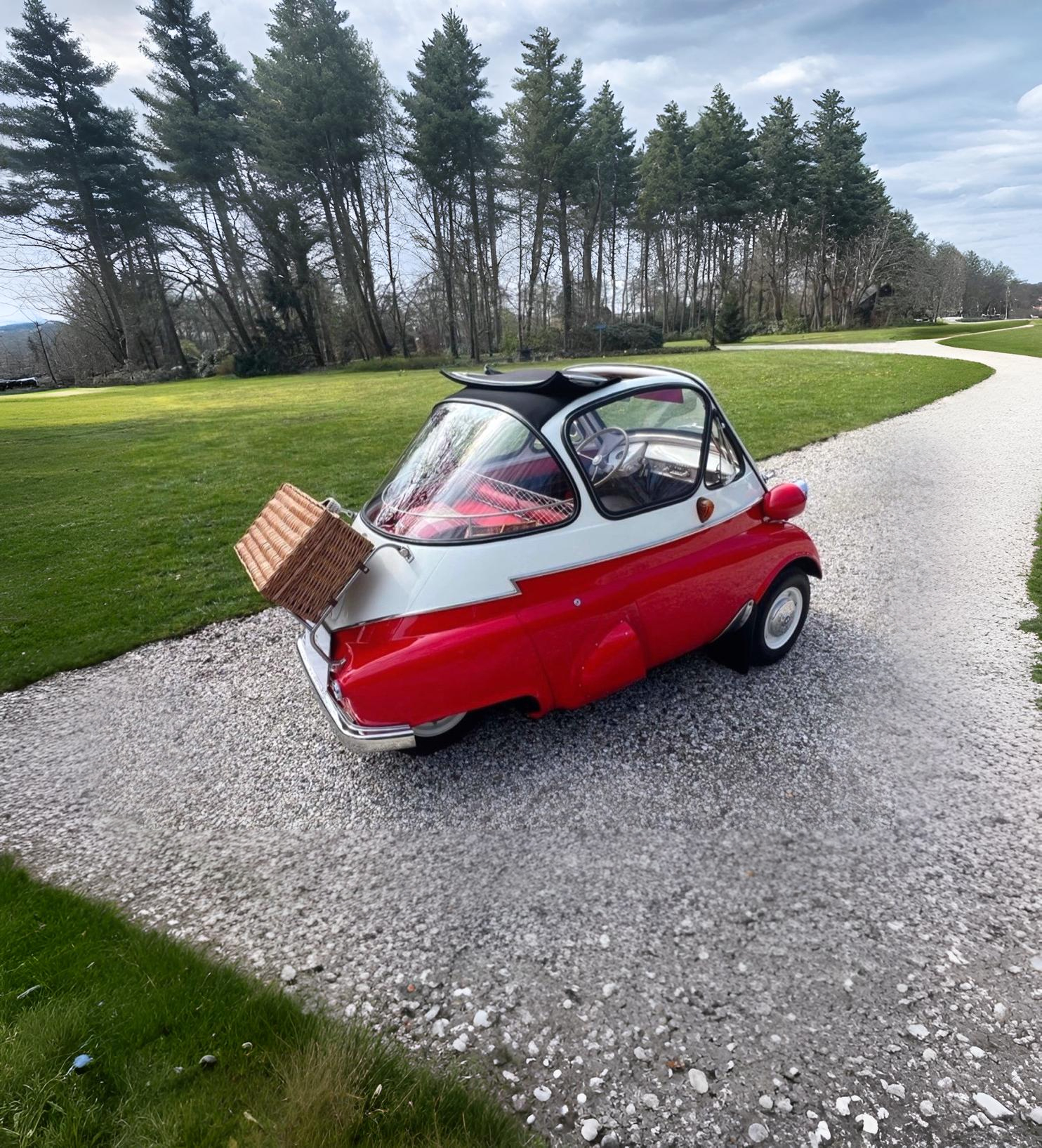 Small vintage red and white microcar with a wicker basket mounted on the rear, parked on a gravel path surrounded by green grass and trees.