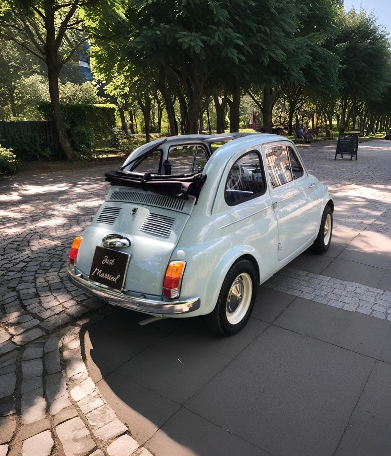 Light blue vintage car with a folded black soft top and a 'Just Married' sign on the rear, parked on a cobblestone and tiled area shaded by trees.