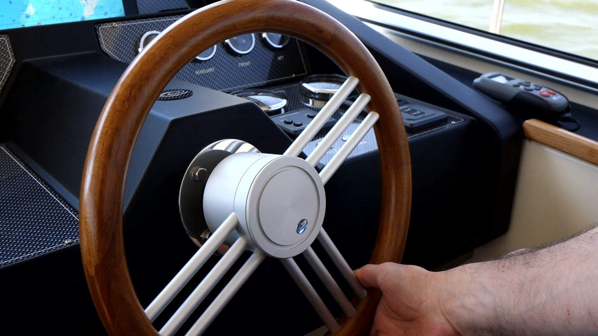Hand gripping a wooden and metal boat steering wheel with instruments and a remote control on the dashboard.