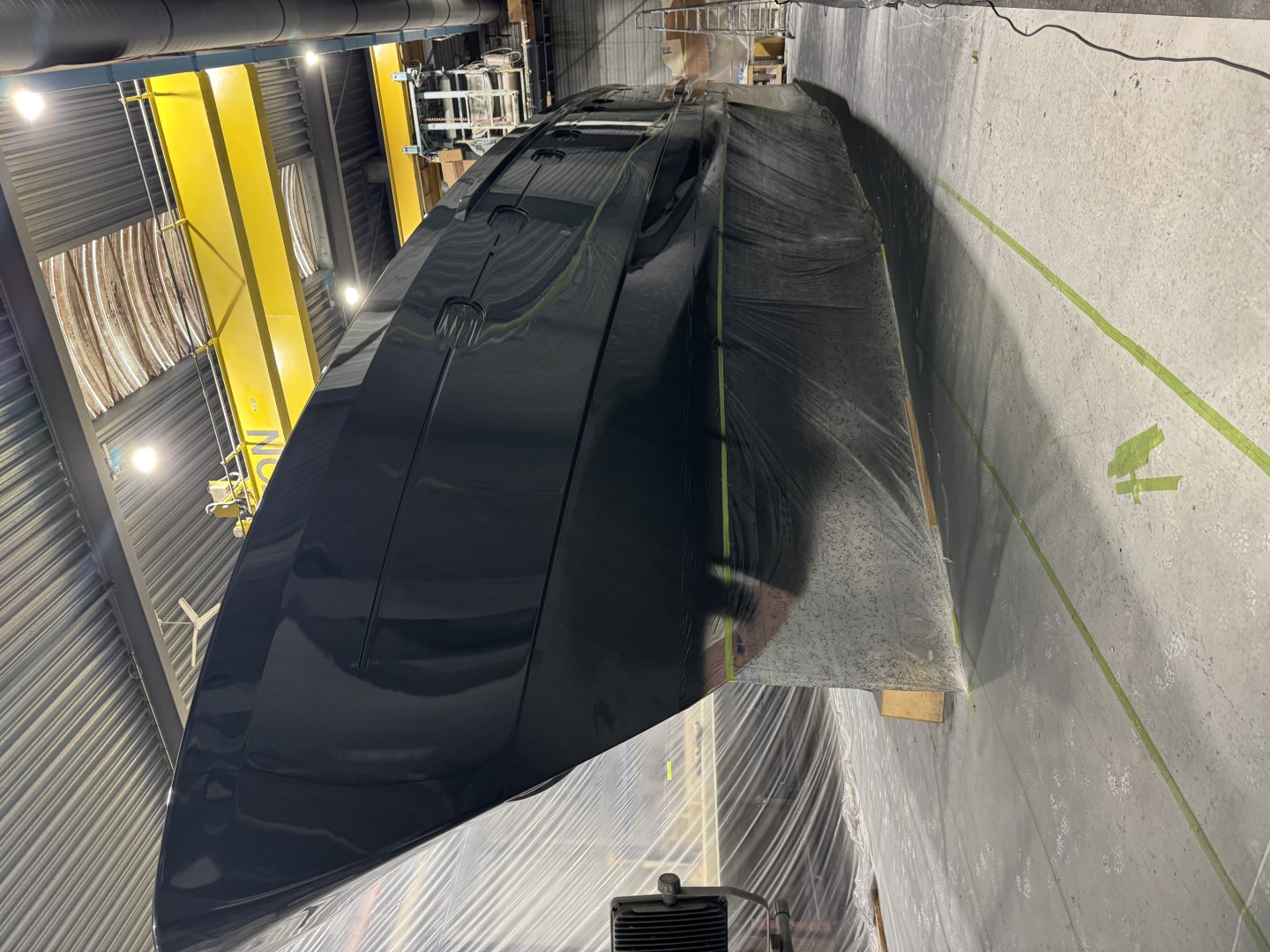 Shiny black boat hull inside an industrial workshop with overhead crane and plastic sheeting covering the floor and walls.