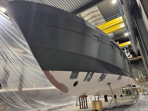 A large boat hull in a workshop being painted with black and white primer, supported by stands and surrounded by plastic sheeting.