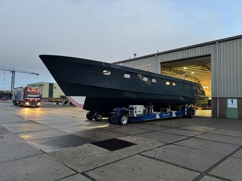 Large black yacht hull on a blue transport vehicle outside a warehouse with its lights on.