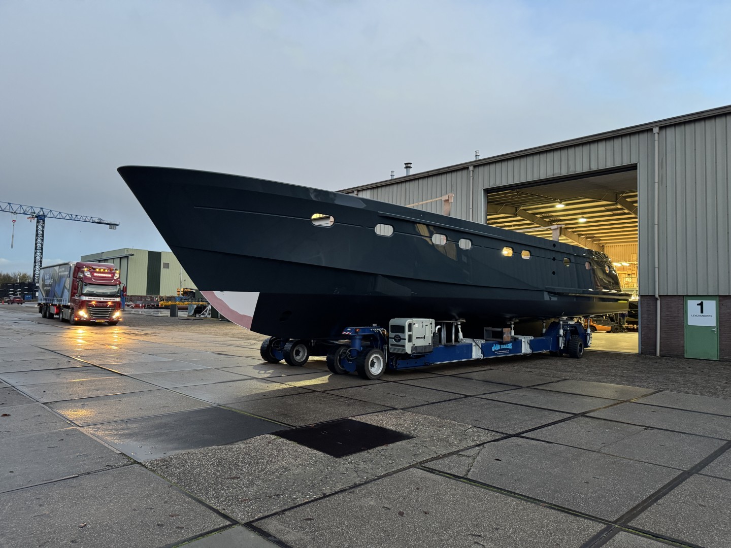 Large black yacht hull on a blue transport vehicle outside a warehouse with its lights on.