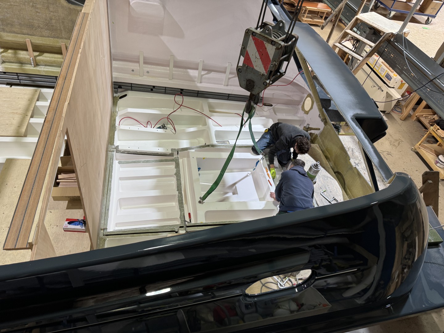 Two workers installing or assembling a large white molded tank or compartment inside a boat hull using a crane in a workshop.