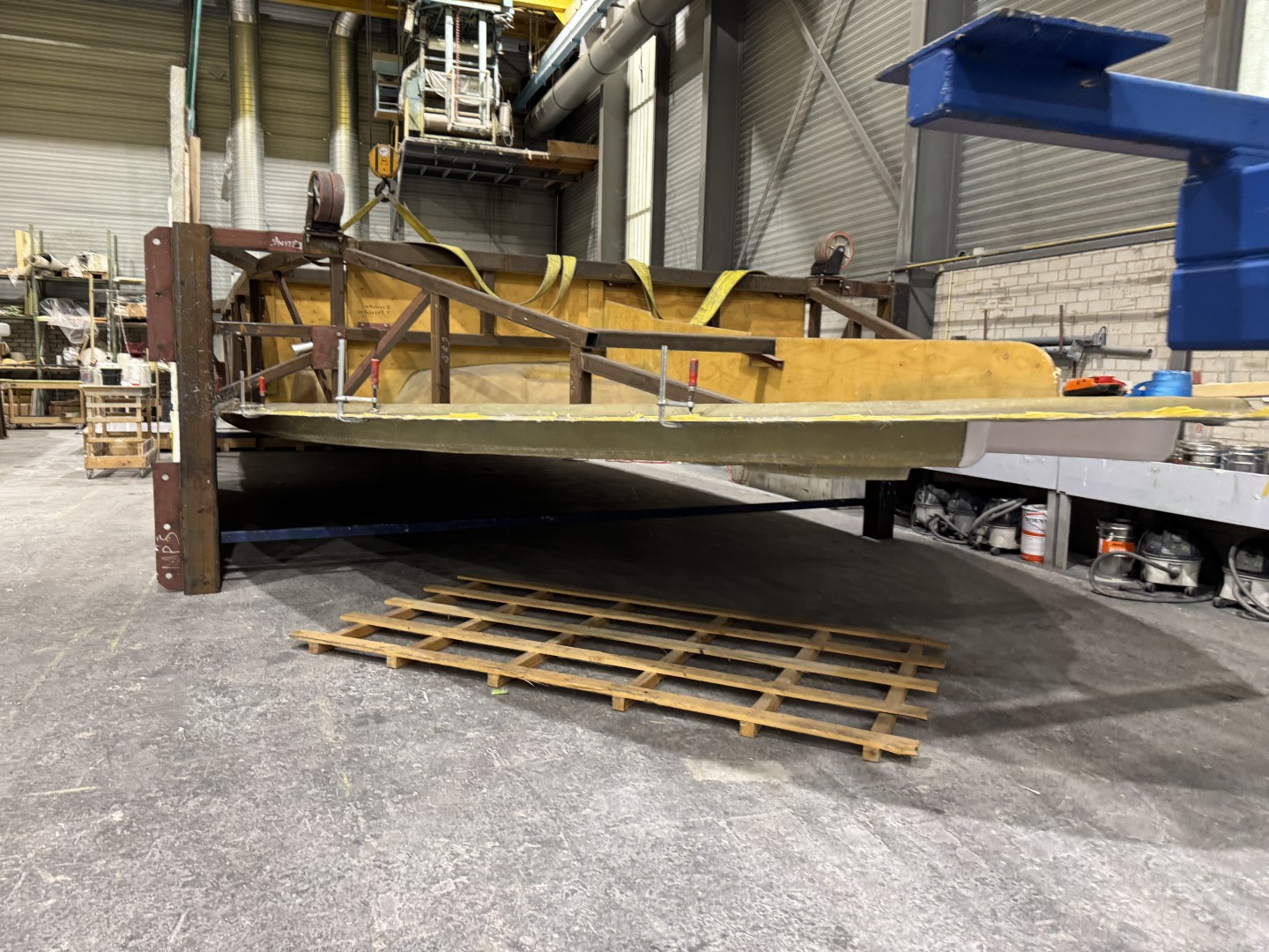Large wooden and metal frame structure suspended in a workshop with industrial equipment and shelving in the background.