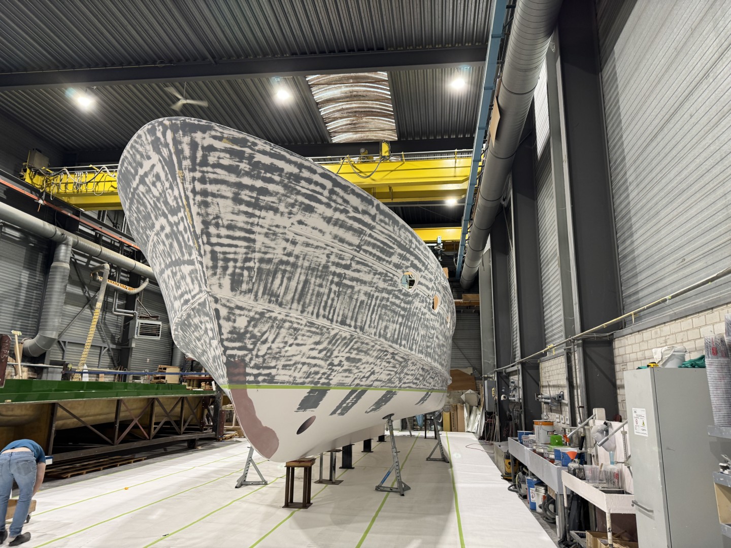 Large boat hull undergoing sanding and preparation inside a spacious industrial workshop with tools and equipment along the walls.