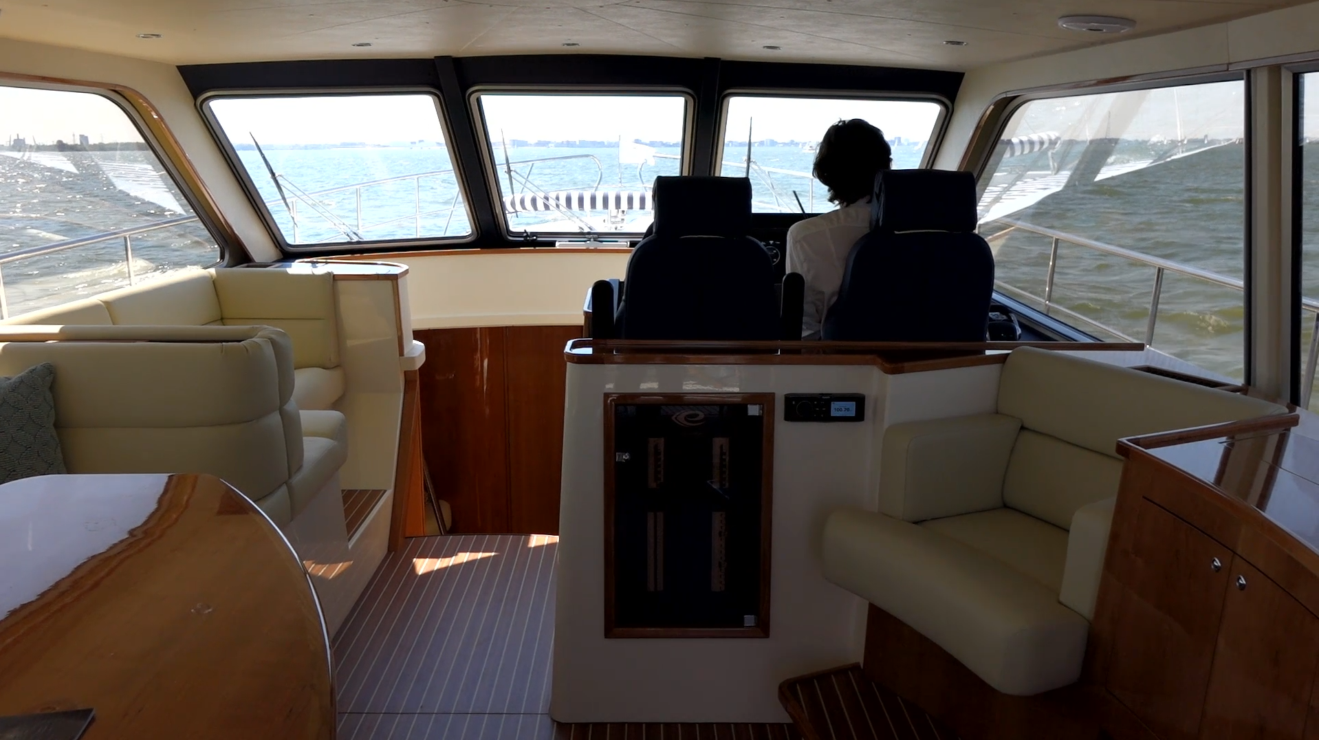 Interior of a boat cabin with beige leather seating, wooden accents, and two people seated at the helm facing forward toward the water.