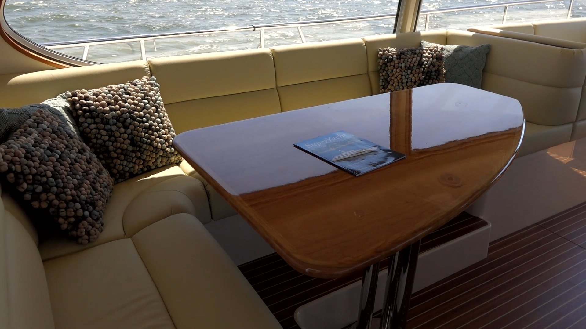 Interior seating area on a yacht with beige cushions, textured throw pillows, and a polished wooden table with a SuperYacht magazine on it, overlooking water.