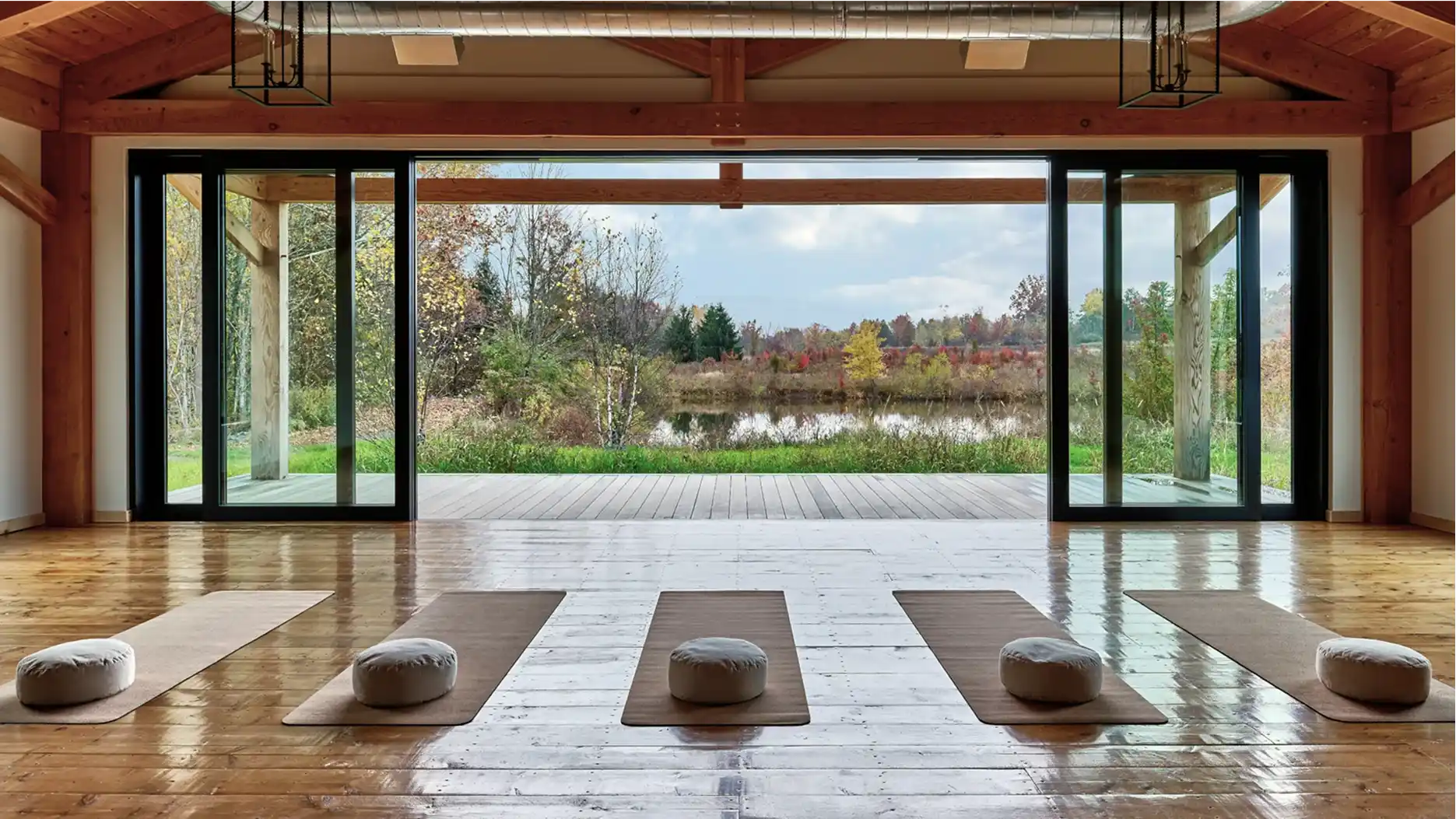 A photo of an elegant yoga studio interior with a group of yoga mats laid out on the floor and headpillows at the end of each. The studio looks out at a beautiful nature view with colorful fall foliage and a pristine natural pond several years away.