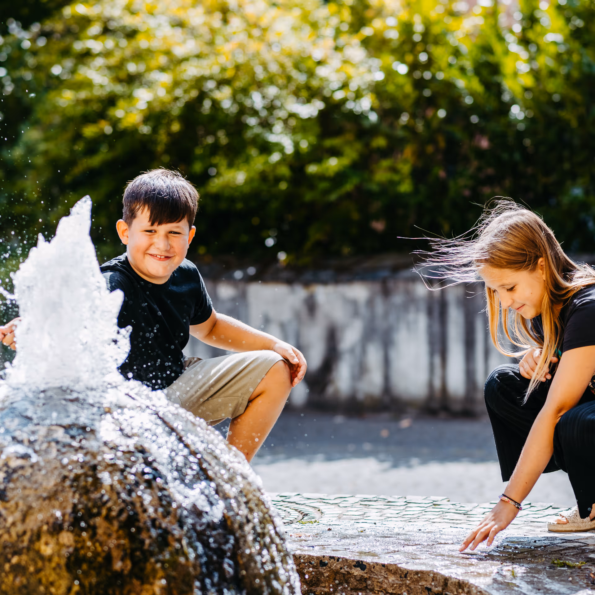 Zwei Kinder spielen fröhlich mit Wasser an einem Brunnen im Freien bei sonnigem Wetter.