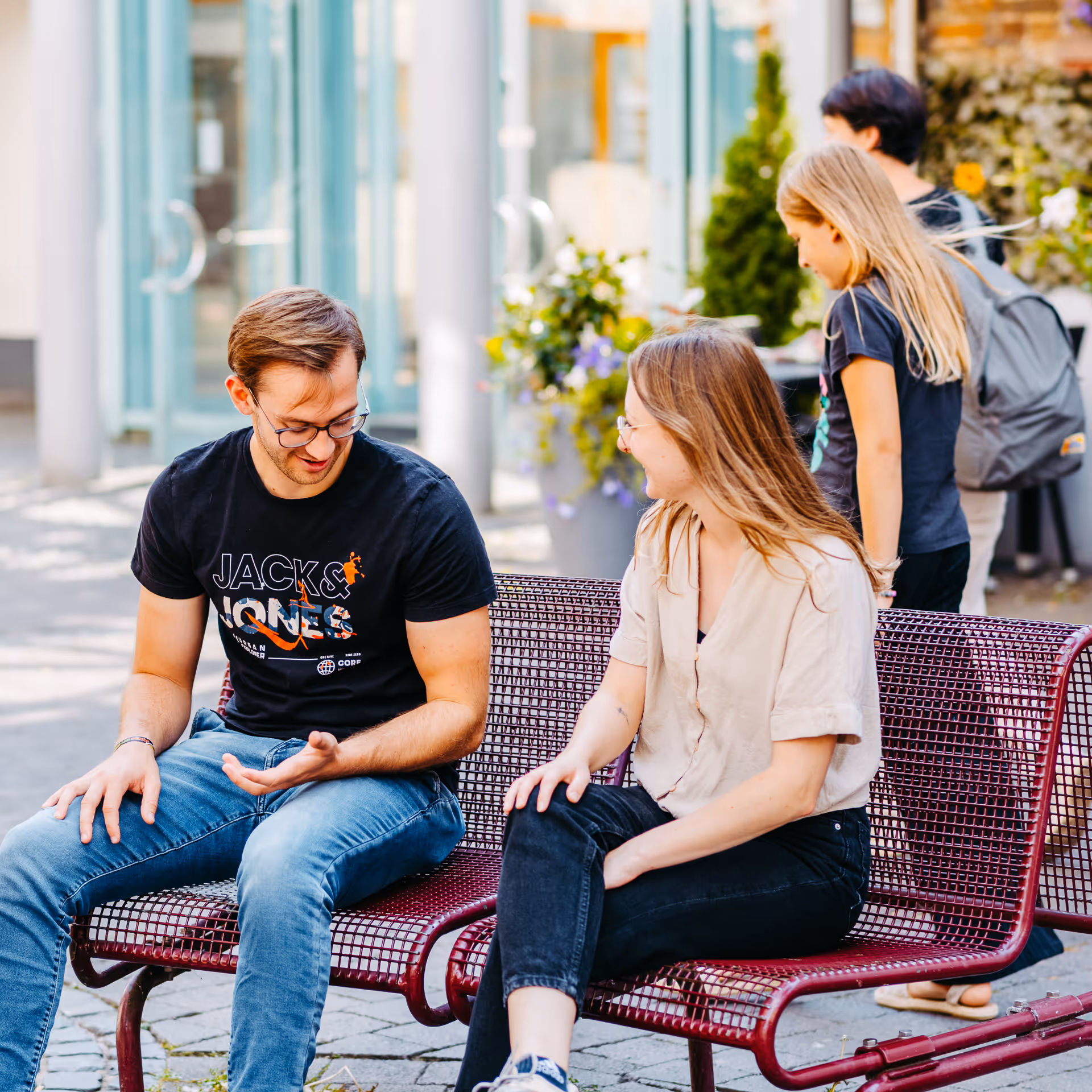 Zwei junge Erwachsene sitzen und unterhalten sich auf einer roten Metallbank in einer städtischen Straße, im Hintergrund gehen zwei weitere Personen vorbei.