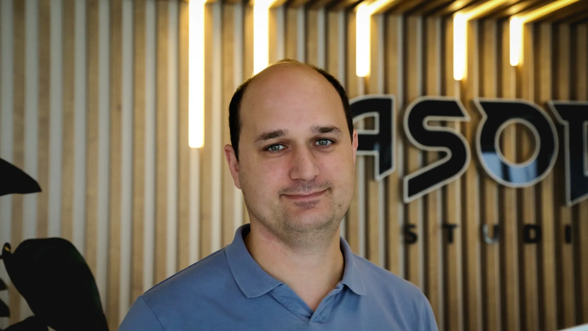 Portrait d’un membre d’Asobo Studio devant le logo de l’entreprise, dans un environnement moderne avec éclairage vertical et fond en bois clair.