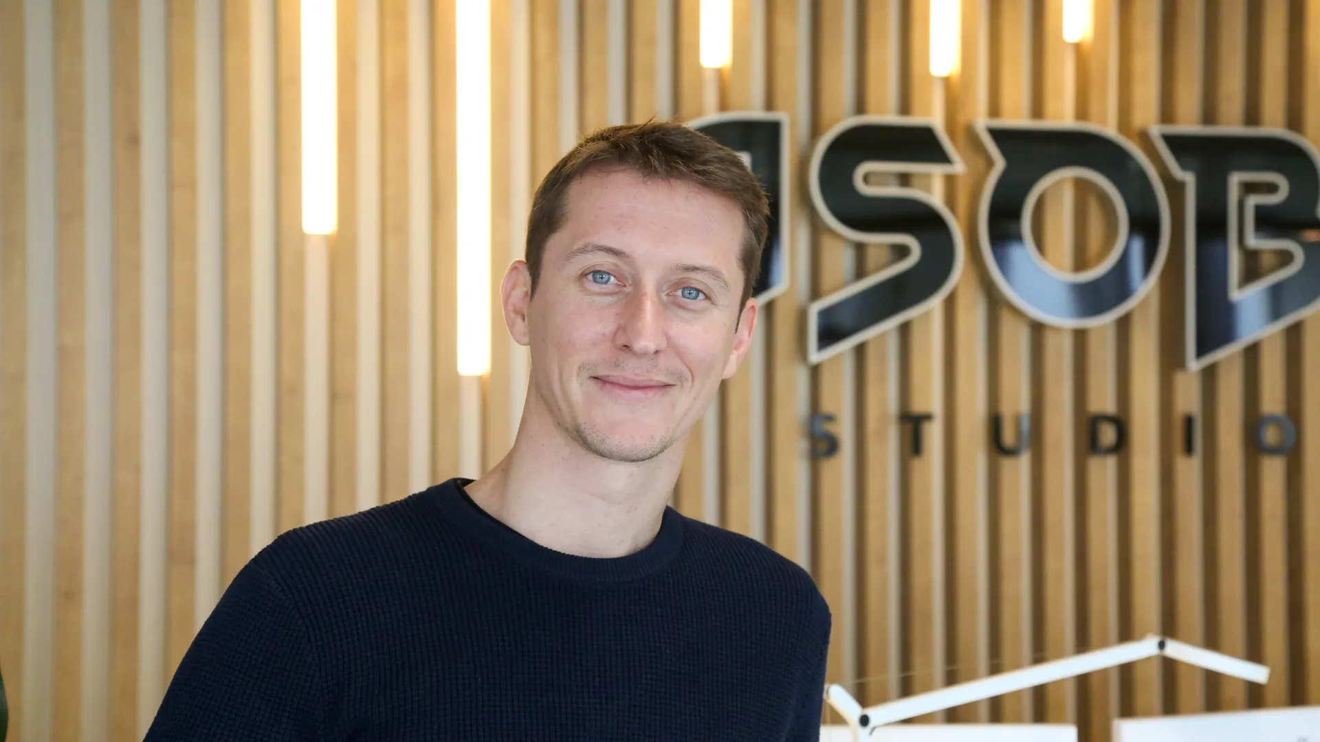 Portrait d’un membre d’Asobo Studio devant le logo de l’entreprise, dans un environnement moderne avec éclairage vertical et fond en bois clair.