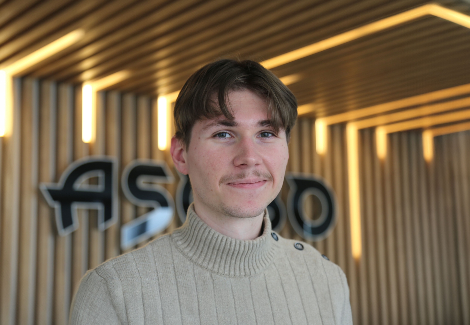 Portrait d’un membre d’Asobo Studio devant le logo de l’entreprise, dans un environnement moderne avec éclairage vertical et fond en bois clair.
