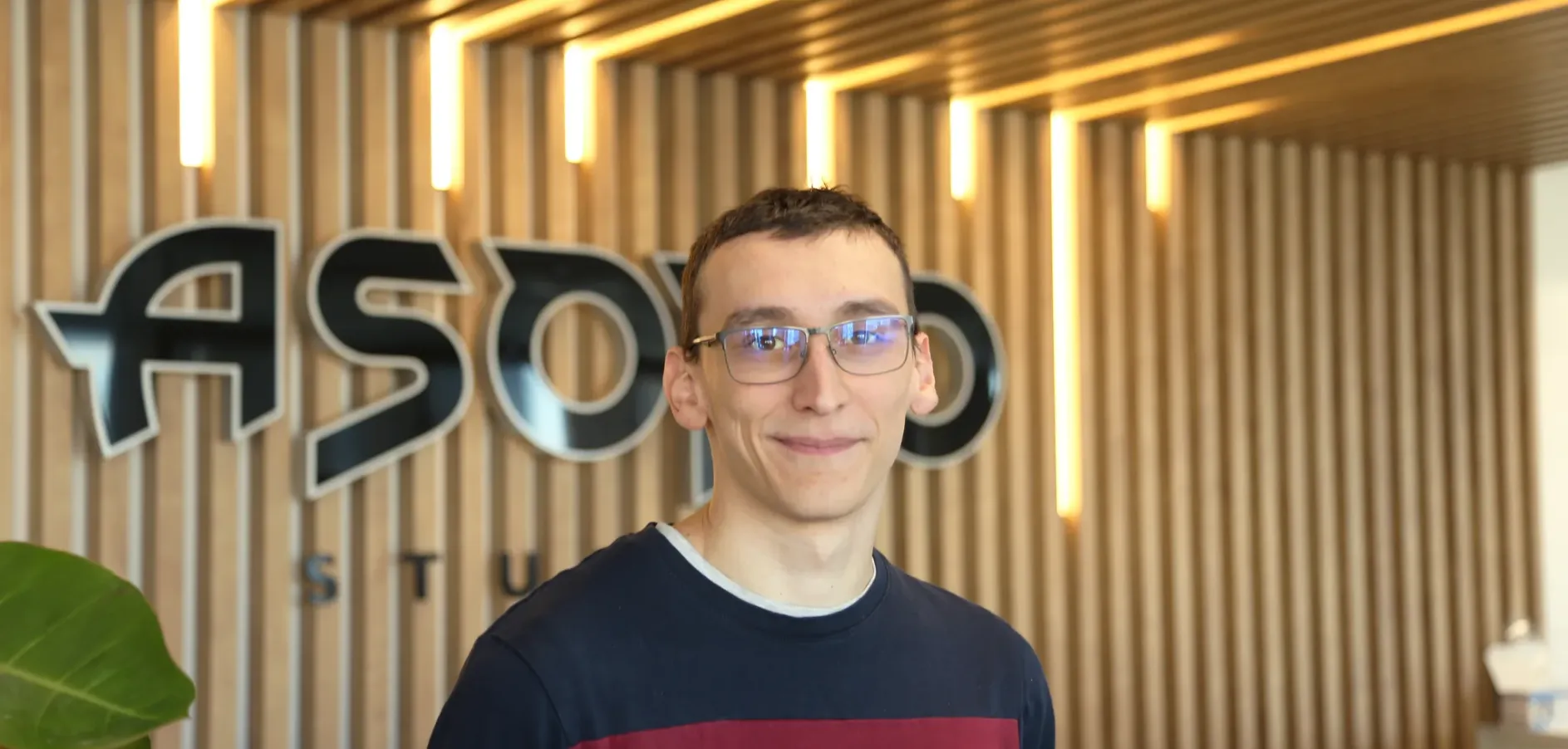 Portrait d’un membre d’Asobo Studio devant le logo de l’entreprise, dans un environnement moderne avec éclairage vertical et fond en bois clair.