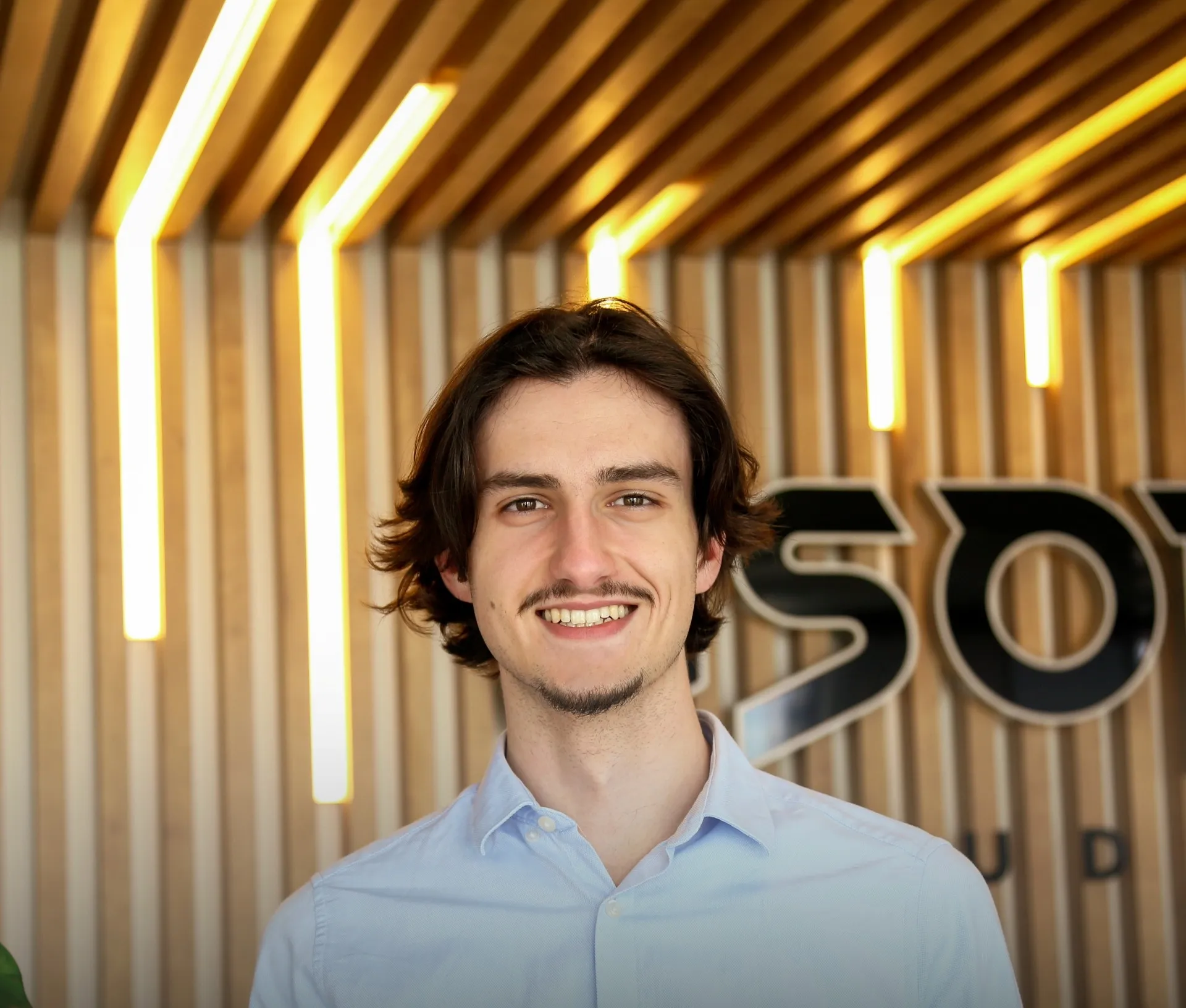 Portrait d’un membre d’Asobo Studio devant le logo de l’entreprise, dans un environnement moderne avec éclairage vertical et fond en bois clair.