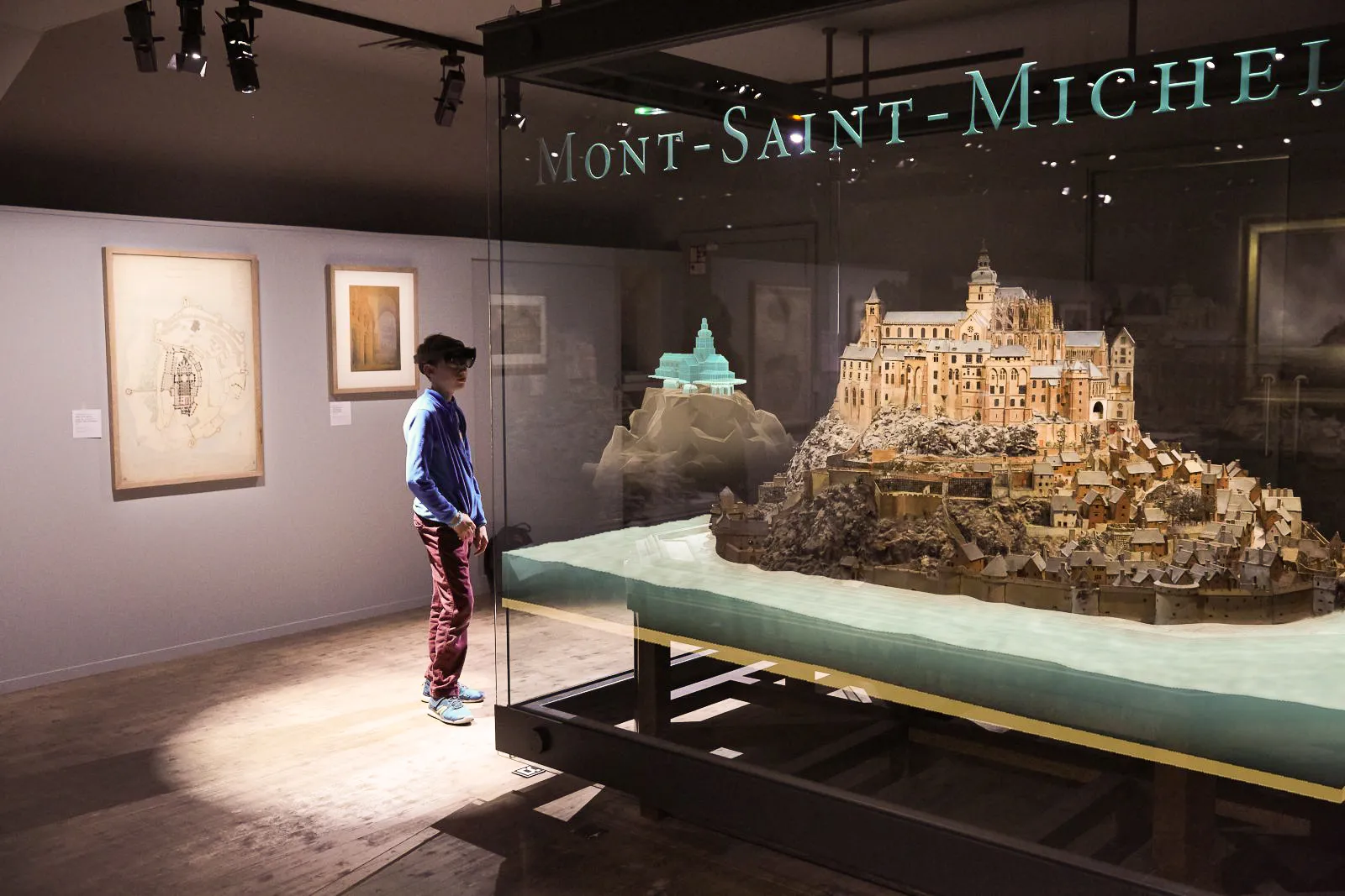 Jeune visiteur utilisant un casque de réalité mixte HoloLens pour explorer une maquette interactive du Mont-Saint-Michel au musée, combinant patrimoine historique et technologie immersive