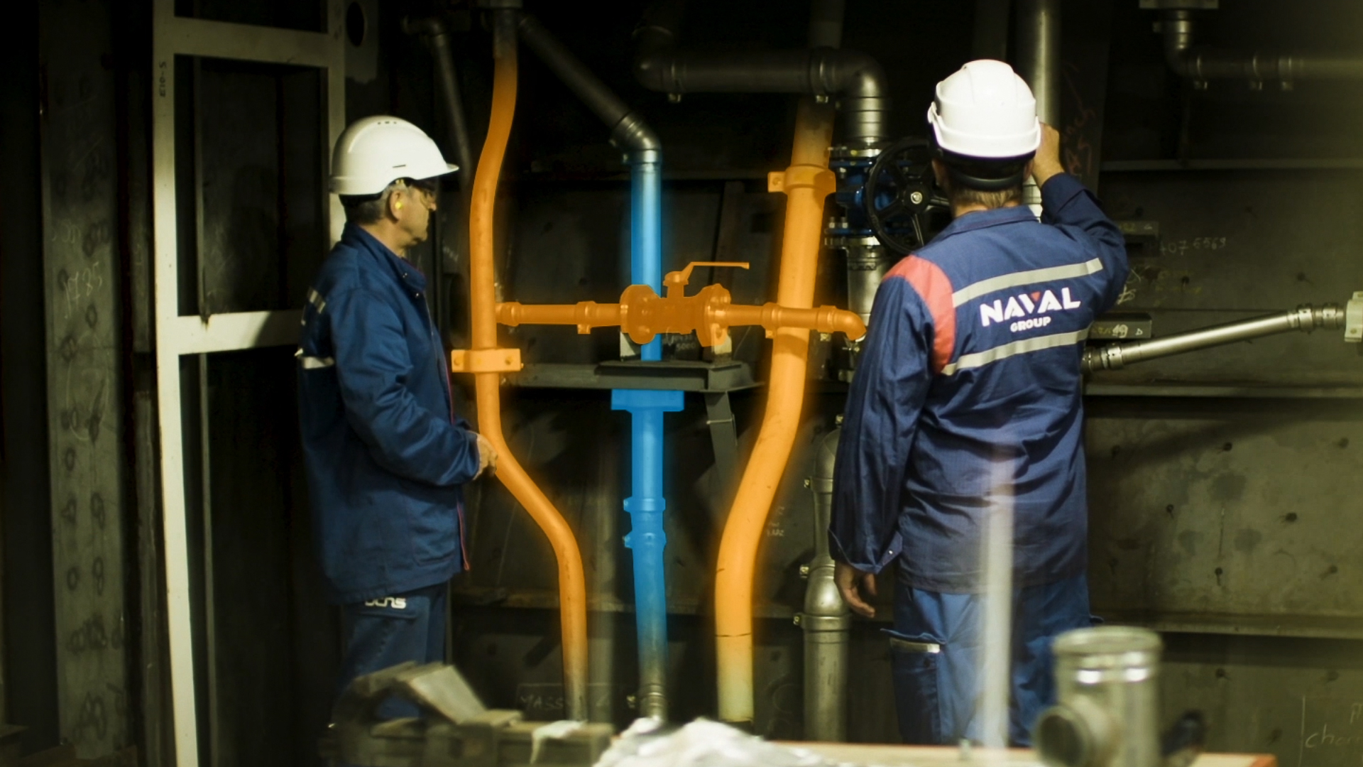 Two Naval Group technicians equipped with safety helmets in front of a pipe network, visualizing colored pipes and valves in augmented reality to optimize maintenance and industrial tracking.