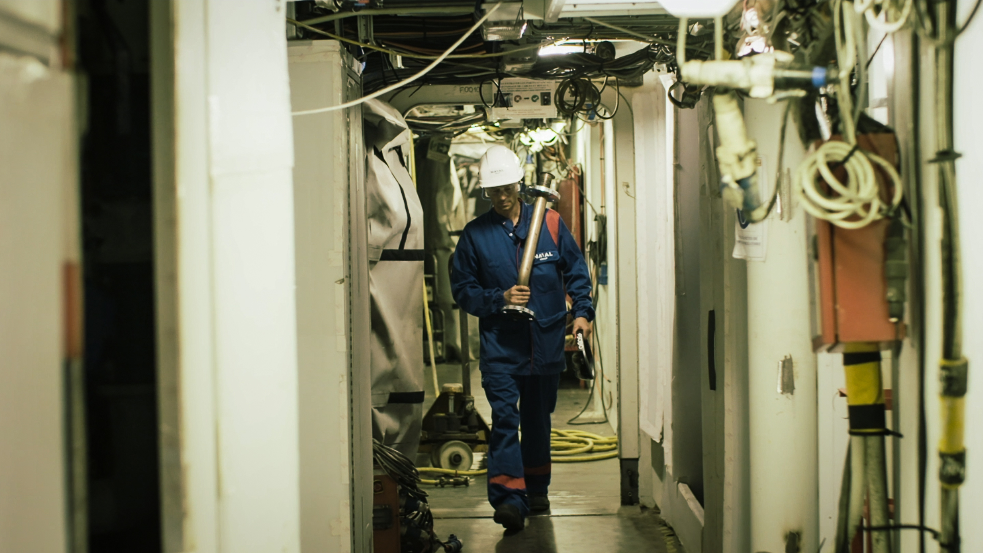 A technician in a blue suit and Naval Group safety helmet carries a metal part in a narrow technical corridor of a ship with his piece of pipes and his HoloLens helmet, illustrating the challenges of maintenance and industrial interventions in a confined environment.