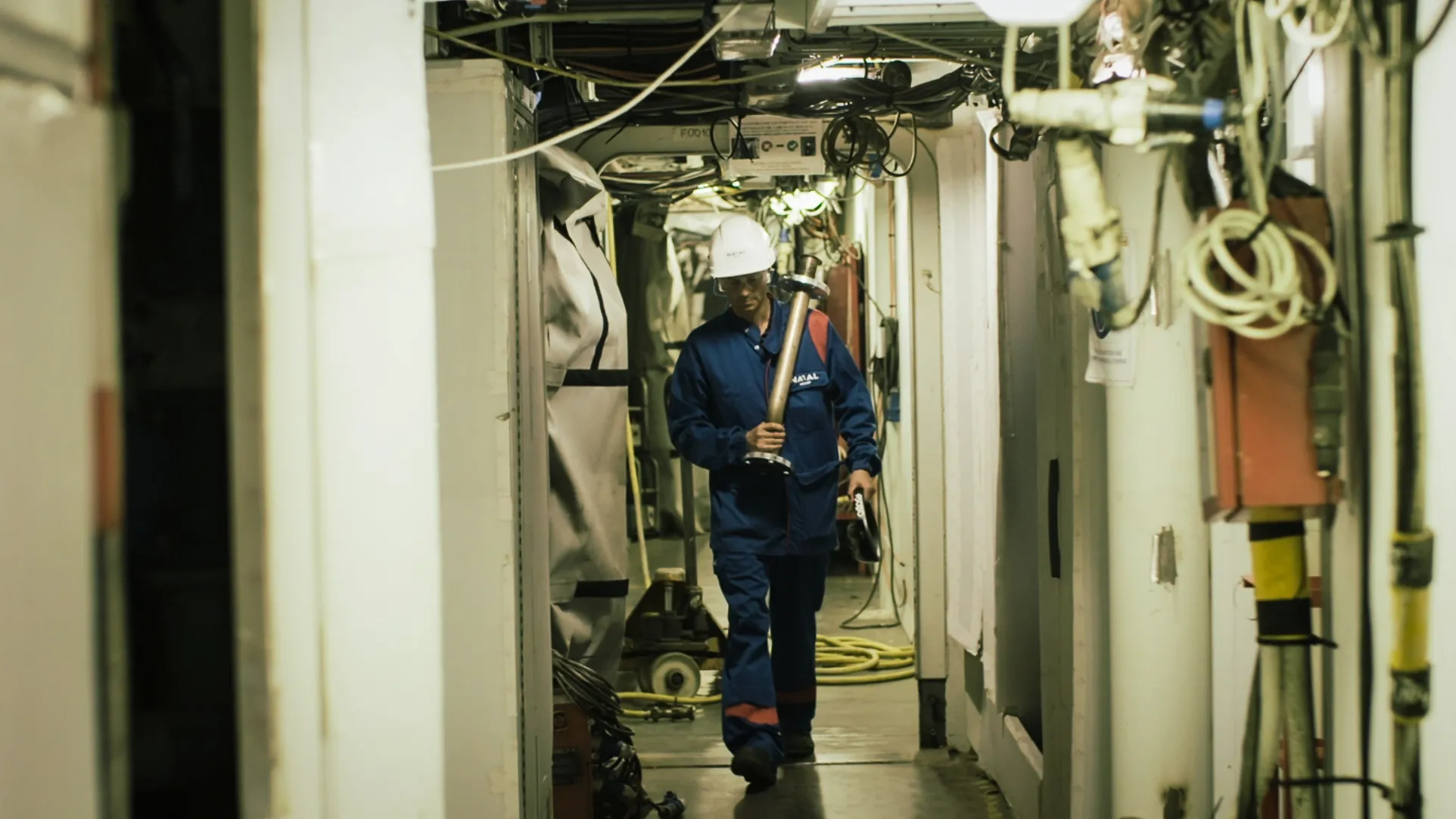 Un technicien en combinaison bleue et casque de sécurité Naval Group transporte une pièce métallique dans un couloir technique étroit d’un navire avec sa pièce de tuyaux et son casque HoloLens, illustrant les défis de la maintenance et des interventions industrielles en milieu confiné.
